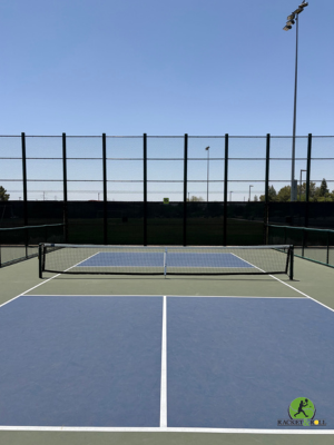 Outdoor tennis court with a net, blue playing surface, white boundary lines, and tall surrounding fence under a clear sky.
