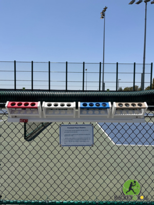 Color-coded ball holders mounted on a fence above a tennis court, with a sign and tennis logo visible.